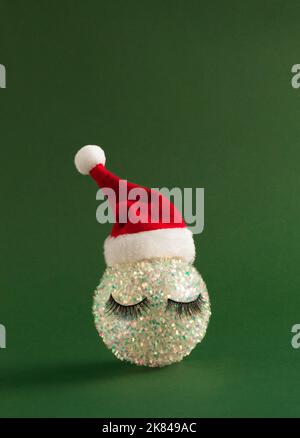 Bianco brillante Natale babble con ciglia e cappello Babbo Natale su sfondo verde audace. Felice anno nuovo sfondo. Foto Stock