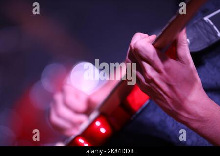 Primo piano della mano e delle dita che suonano musica su una chitarra elettrica rossa brillante. Il fret di strumming della mano con uno sfondo deliberatamente sfocato del riflettore Foto Stock