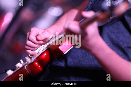 Primo piano della mano e delle dita che suonano musica su una chitarra elettrica rossa brillante. Il fret di strumming della mano con uno sfondo deliberatamente sfocato del riflettore Foto Stock