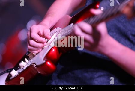 Primo piano della mano e delle dita che suonano musica su una chitarra elettrica rossa brillante. Il fret di strumming della mano con uno sfondo deliberatamente sfocato del riflettore Foto Stock