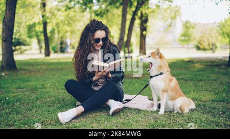 Allegra studentessa sta leggendo un libro nel parco seduto sul prato e carezzando bel cane, l'animale domestico è ragazza sbadiglio è ridere. Foto Stock