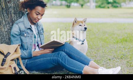 Ritratto di grazioso libro afro-americano studente di lettura nel parco seduto su erba con il suo cane obbediente vicino. Stile di vita moderno, gioventù, natura e animali concetto. Foto Stock