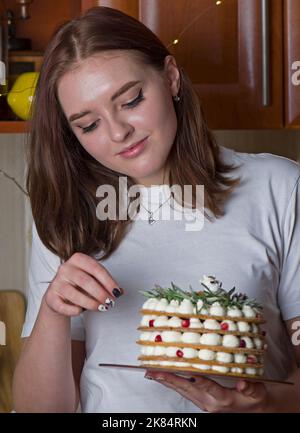 Una bella ragazza felice in una T-shirt bianca, si alza a casa, tiene una torta di Capodanno festosa su un piatto Foto Stock