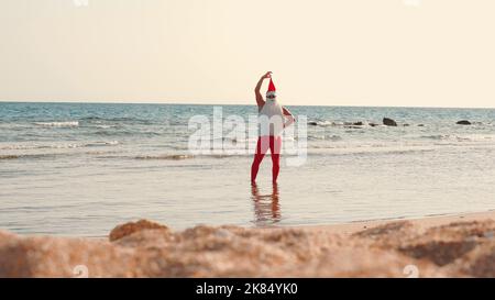 Babbo Natale si bagna il sole. Babbo Natale divertendosi. Babbo Natale divertente, in occhiali da sole, in piedi in acqua di mare sulla spiaggia. Vacanze estive a Babbo Natale. Foto di alta qualità Foto Stock