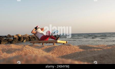 Babbo Natale si bagna il sole. Divertente Babbo Natale, in occhiali da sole e pinne, è seduto sul lettino, sulla spiaggia sul mare e ascoltando musica con un piccolo altoparlante musicale in mano, rilassante. vacanze estive babbo natale, in riva al mare. Foto di alta qualità Foto Stock