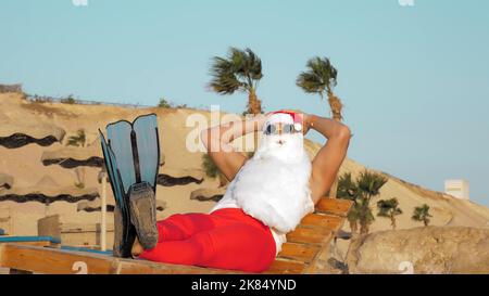 Vacanze estive a Babbo Natale. Babbo Natale si bagna il sole. Fundy Babbo Natale, in occhiali da sole e pinne, è seduta, rilassarsi sul lettino, sulla spiaggia sul mare, sullo sfondo di palme. Foto di alta qualità Foto Stock