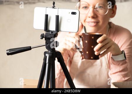 una ragazza con una tazza di caffè solleva il suo telefono su un treppiede registra una video lezione online, formazione online e conferenza in quarantena Foto Stock