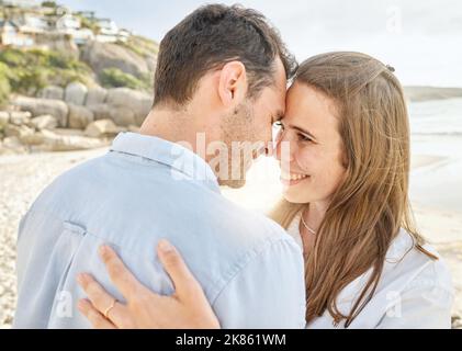 Coppia, amore e abbraccio su una spiaggia insieme per un fidanzato luna di miele o anniversario dal mare. Giovane uomo e donna sorridono sentendosi felici e romantici Foto Stock