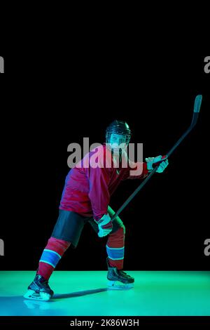 Lancio potente. Giocatore di hockey su ghiaccio maschile in uniforme sportiva protettiva in azione su sfondo scuro in luce al neon. Sport, potenza, sfide Foto Stock