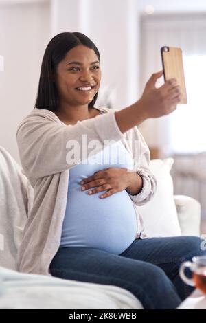 La gravidanza è un viaggio bellissimo, cattura ogni momento di esso. Una donna incinta che prende un selfie mentre si siede sul divano a casa. Foto Stock