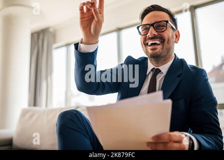Elegante uomo d'affari in tuta, con la mano in su, con un documento, seduto sul divano in ufficio. Foto Stock