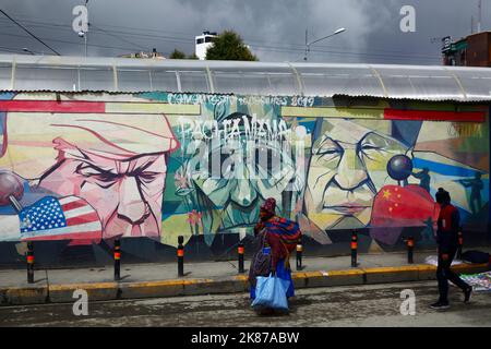 Signora Aymara che cammina davanti al murale mostrando Pachamama (Madre Terra) come una donna anziana triste fiancheggiata da Donald Trump e Xi Jinping, la Ceja, El Alto, Bolivia Foto Stock