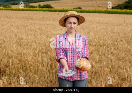 Giovane agricoltore che tiene dollari e pane in campo di grano dorato. Profitto dall'agricoltura durante la stagione di raccolta in estate. Foto Stock