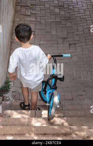 Vista posteriore del bambino che trasporta la bicicletta blu di equilibrio mentre muovendosi giù sulla scala. Foto Stock