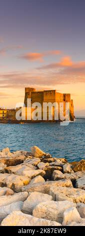 Napoli, Italia. Castel dell'Ovo con rocce frangiflutti in primo piano e con un bel cielo tramonto. Banner verticale. Foto Stock