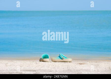 Regione di Krasnodar. Russia. Luglio 19, 2022. Un paio di sandali Crocs blu su una spiaggia sabbiosa con sfondo mare sfocato. Scarpe da spiaggia in riva al mare in una giornata di sole estate. Concetto di vacanza in spiaggia. Foto Stock