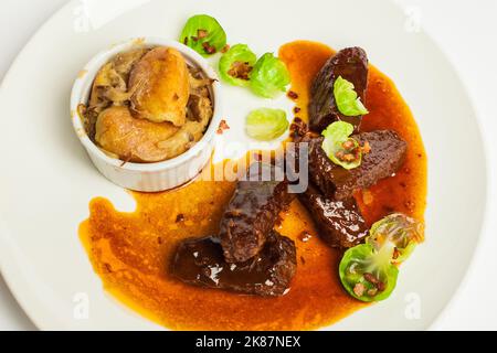 Guance di vitello in salsa rossa con verde e patate primo piano su un piatto bianco su fondo bianco Foto Stock