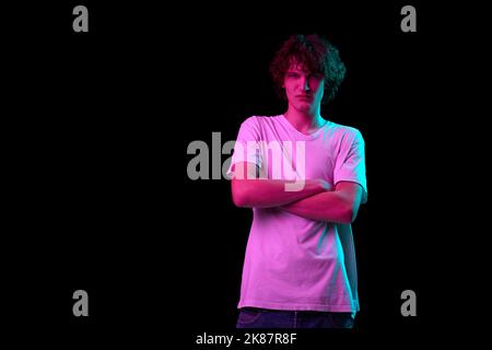 Giovane arrabbiato in piedi con braccia incrociate isolato su sfondo scuro in luce al neon viola. Emozioni, gioventù, vendite, pubblicità Foto Stock