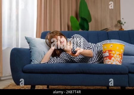 Una giovane donna depressa si trova sul divano e cambia canale sul televisore Foto Stock