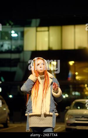 Content donna che ascolta musica in città di notte. Giovane donna positiva con lunghi dreadlock arancioni e in giacca denim ascoltando musica in headpho Foto Stock