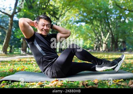 Ritratto di un atleta nel parco, un uomo asiatico guarda la macchina fotografica e sorride, un uomo in una tuta sportiva fa esercizi fisici su un materassino sportivo. Foto Stock