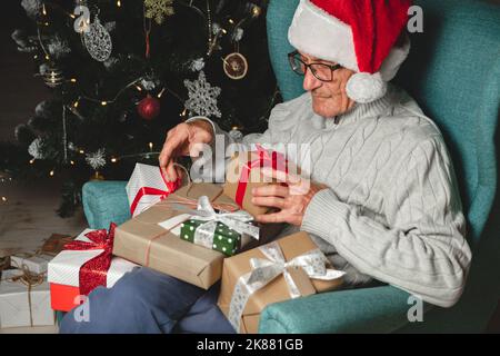 Anziano in un cappello di babbo natale con i lotti dei regali di Natale siede io Foto Stock