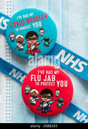 Colorati badge Covid 19 Booster e flu jab che si trovano sulla parte superiore di una maschera chirurgica e legati a un cordone NHS Foto Stock