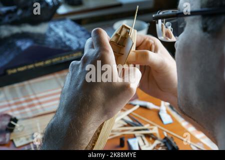 Mani di uomo incollando con colla i particolari del compensato per modello di nave, tenendo con le dita. Processo di costruzione di navi giocattolo, hobby, artigianato. Tabella con Foto Stock