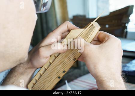 Mani di uomo incollando con colla i particolari del compensato per modello di nave, tenendo con le dita. Processo di costruzione di navi giocattolo, hobby, artigianato. Tabella con Foto Stock