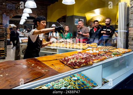 Una lavoratrice di ristoranti affetta fette di pizza per i clienti dietro una vetrina in una piccola paninoteca e pizzeria di Bergamo Italia. Foto Stock