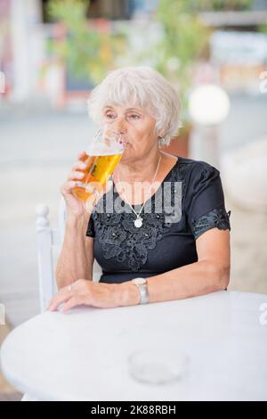 Donna matura seduta a un tavolo in un bar estivo e bere birra Foto Stock