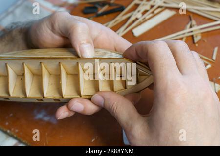 Mani di uomo incollando con colla i particolari del compensato per modello di nave, tenendo con le dita. Processo di costruzione di navi giocattolo, hobby, artigianato. Tabella con Foto Stock