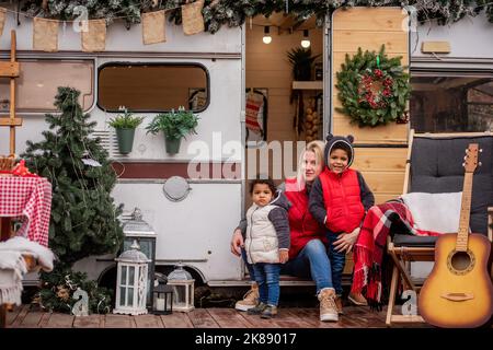 La madre caucasica gioca divertendosi con i bambini afroamericani sulla casa del rimorchio della porta d'ingresso. Giovane donna abbraccia bacia figlio, figlia. L'elogio di famiglia Foto Stock