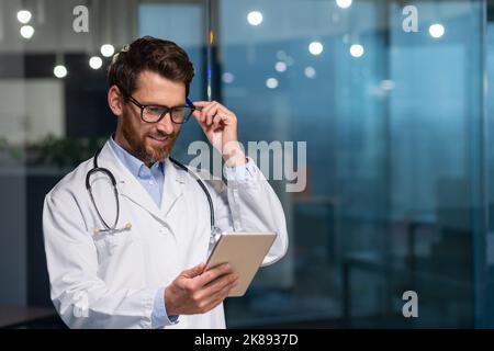 Allegro e sorridente medico senior in occhiali e abito medico leggere messaggio da tablet computer, uomo che lavora all'interno di clinica moderna Foto Stock