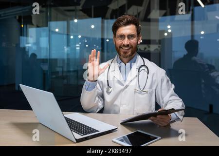 Ritratto di un medico maturo di successo all'interno di una clinica moderna, un uomo che lavora con un computer portatile guardando la macchina fotografica e sorridente gesto di saluto con la mano ondulata Foto Stock