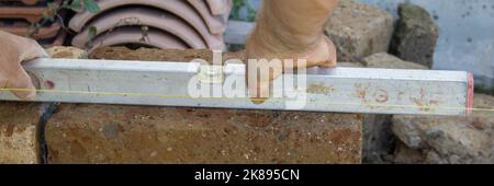 Immagine delle mani di un muratore che controlla la regolarità del piano del muro di tufo che sta costruendo con un livello. Banner orizzontale Foto Stock