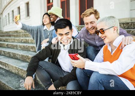 Gruppo di giovani amici multirazziali seduti all'aperto con i loro telefoni Foto Stock