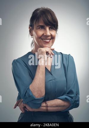 Sognare e pensare a qualcosa di nuovo. Studio shot di una donna anziana che posa con la mano sul mento su uno sfondo grigio. Foto Stock