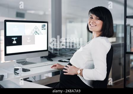 Ho una mini-collega nella mia pancia. Una donna d'affari incinta attraente seduta nel suo ufficio e usando il suo computer mentre tiene la pancia. Foto Stock