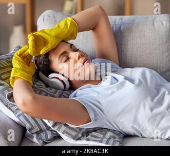 Divano, cuffie e donna più pulita dormire, rilassarsi e ascoltare la musica dopo aver pulito il lavoro nel salotto di casa. Calmo, stanco e ragazza fatica su un Foto Stock