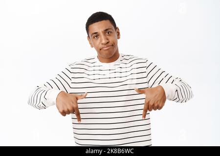 Un ragazzo afro-americano deluso, che guarda scettico alla telecamera, punta le dita verso il basso, in piedi su sfondo bianco Foto Stock