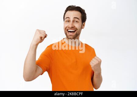 Ragazzo bearded entusiasta mostrando il suo sostegno, allegra, alzando il pugno e celebrando, trionfando, in piedi su sfondo bianco Foto Stock