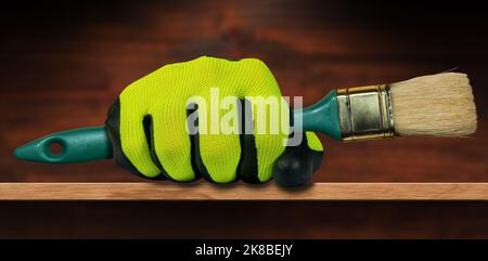 Operatore manuale con guanti da lavoro di protezione verdi e neri che regge un pennello usato con manico verde, su un banco di lavoro in legno con spazio per copiare. Foto Stock