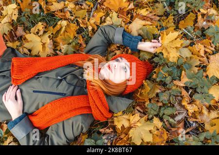 Donna sognante con foglie gialle autunnali in mano in cappello a maglia arancione e scarflatto sdraiato sulle foglie nel parco Foto Stock