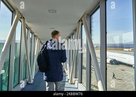 Vista posteriore dell'irriconoscibile viaggiatore maschio che cammina lungo l'aeroporto con zaino. Vista posteriore di un uomo che passa dal corridoio del terminal sulla via del volo Foto Stock