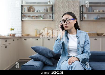 Preoccupata e confusa giovane donna asiatica seduta sul divano a casa e chiamata servizio di emergenza su cellulare. Parla al telefono, chiede aiuto. Foto Stock