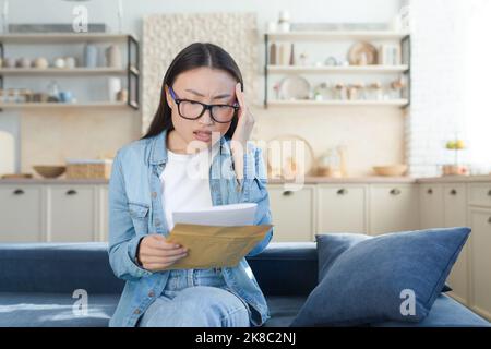 Scioccato e allarmato giovane bella donna asiatica ha ricevuto documenti di divorzio. Seduti a casa sul divano e tenendo una busta con i fogli nelle mani. Tenendo la testa da notizie inaspettate. Foto Stock