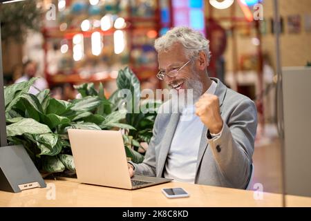 Felice uomo d'affari anziano che utilizza il notebook per celebrare i buoni risultati vincendo online. Foto Stock