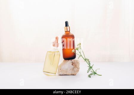 Due bottiglie di olio cosmetico o siero su una pietra e fiore di yarrow su sfondo bianco. Cosmetici naturali, spa e concetto di benessere Foto Stock