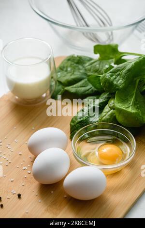 Tre uova bianche di pollo su un tagliere con un mazzo di spinaci freschi. Un uovo crudo in un recipiente di vetro. Un bicchiere di latte sullo sfondo. Foto Stock